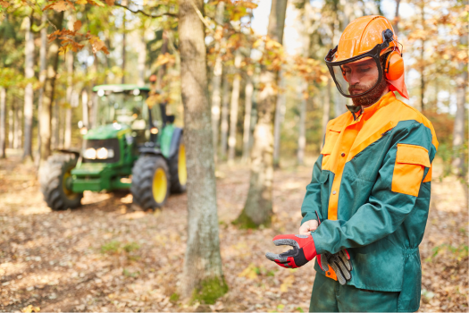 A forestry worker in a safety helmet, face shield, and protective gear putting on gloves in a wooded area with a green tractor in the background.