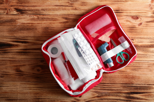 Top-down view of an open red first aid kit on a wooden surface, containing medical supplies like bandages, pills, an inhaler, scissors, and a syringe.