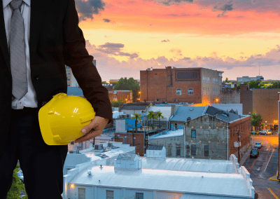 A businessman in a suit holds a yellow hard hat under his arm, standing in front of a city skyline at sunset.