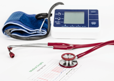 A red stethoscope, a digital blood pressure monitor with a blue cuff, and a medical chart with graphs are arranged on a white surface.