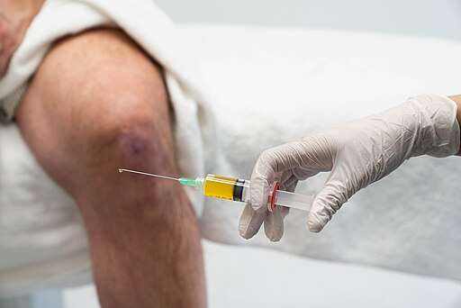 A gloved hand holding a syringe filled with yellow fluid near a patient's knee.