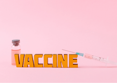 A glass vial with pink liquid, a syringe, and wooden letters spelling "VACCINE" are arranged on a pink background.