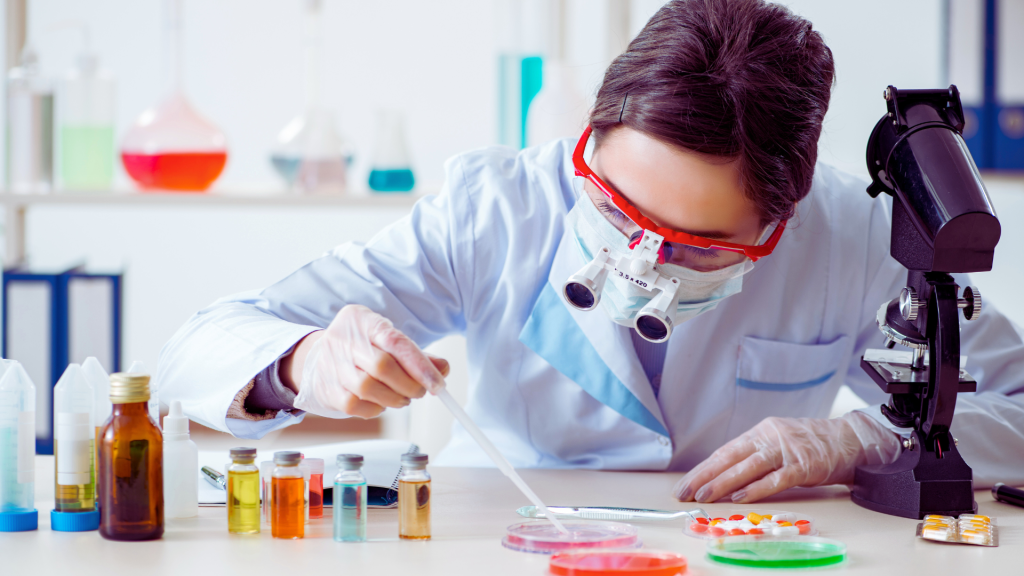 Scientist in a lab setting, wearing protective gear and using a microscope to examine samples in petri dishes and vials.