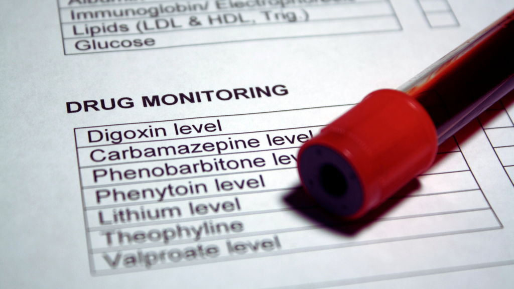 Blood sample vial resting on a "Drug Monitoring" medical form listing tests for various medication levels.