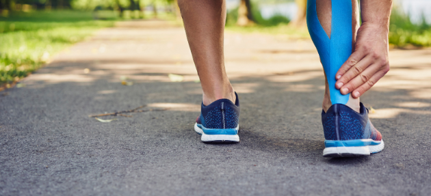 A runner wearing running shoes touches their right heel, which is supported by a strip of blue kinesiology tape running up the calf.