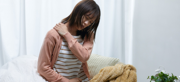 A woman sitting on a bed, clutching her shoulder with a pained expression.