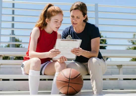 Teenage basketball athlete sitting with her parent on outdoor bleachers reviewing a sports physical form together.