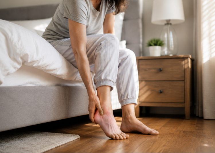 A woman sits on the edge of her bed in the morning, gently holding her heel in pain beside a sunlit bedroom window.