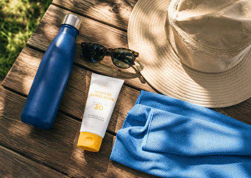 A flat lay of summer heat safety essentials, including a reusable water bottle, sunglasses, sunscreen, a sun hat, and a cooling towel.